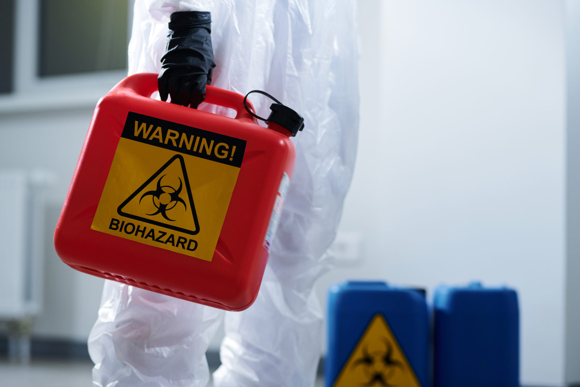 Close-up of unrecognizable laboratory worker in biohazard suit and gloves carrying container with biohazard symbol over corridor